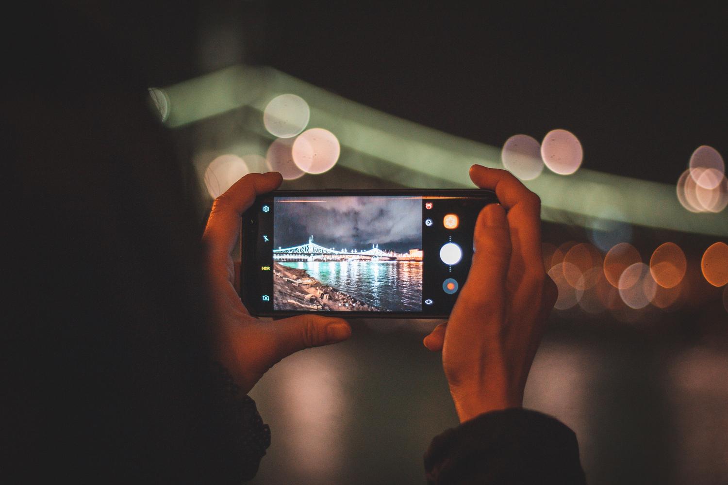 iPhone ekrāna tuvplāns, kas iemūžina apgaismotu pilsētu naktī iPhone ekrāna tuvplāns, kas iemūžina apgaismotu pilsētu naktī ar bokeh efektu, rādot augstas kvalitātes rezultātu.