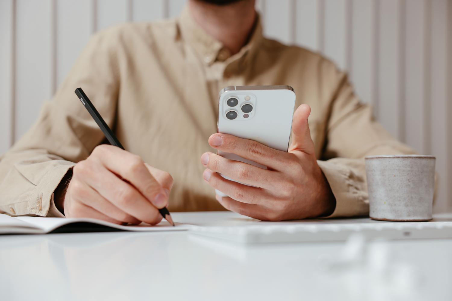 Persona veic piezīmes piezīmju grāmatiņā, pārskatot darba piedāvājumus savā iPhone tālrunī. Persona veic piezīmes piezīmju grāmatiņā, pārskatot darba piedāvājumus savā iPhone tālrunī.