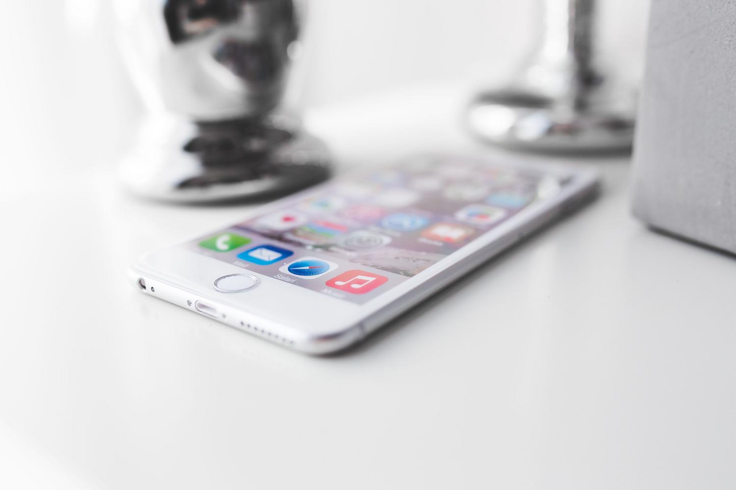 Sudraba iPhone uz balta galda ar ieslēgtu ekrānu, kas attēlo iOS lietotāja interfeisu. Sudraba iPhone uz balta galda ar ieslēgtu ekrānu, kas attēlo iOS lietotāja interfeisu.
