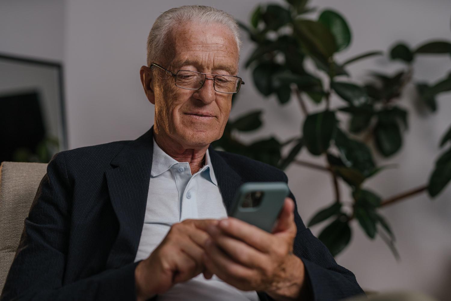 Vecākais vīrietis, kurš smaida, ērti pārlūkojot savu mobilo tālruni mājās. Vecākais vīrietis, kurš smaida, ērti pārlūkojot savu mobilo tālruni mājās.