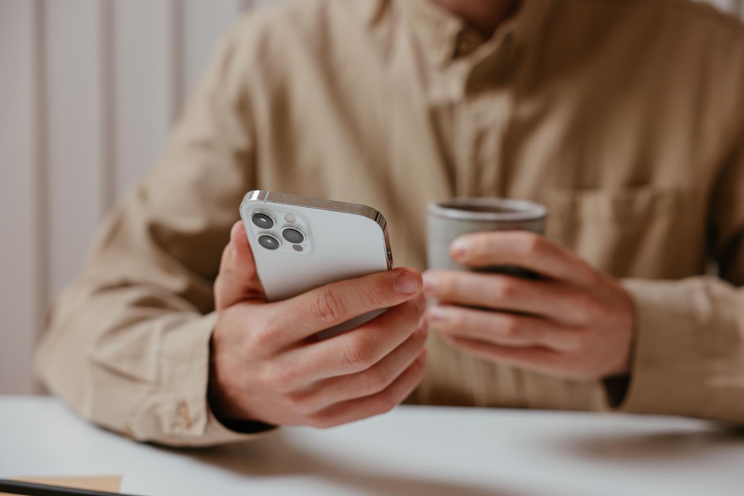 Rokas, kas tur pie kafijas tases moderno iPhone, ilustrē mūsdienu digitālās dienasgrāmatas glabāšanas rituālu Rokas, kas tur pie kafijas tases moderno iPhone, ilustrē mūsdienu digitālās dienasgrāmatas glabāšanas rituālu