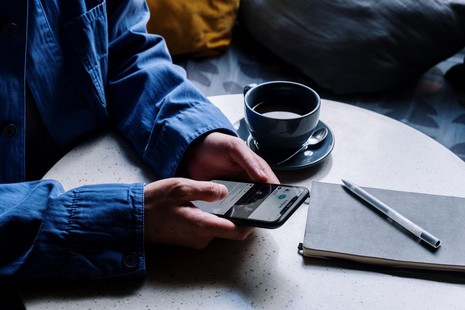 Persona, kas izmanto iPhone tālruni blakus piezīmjdatoram un kafijas tasei nepiespiestā atmosfērā, liekot domāt par pārdomu brīdi Persona, kas izmanto iPhone tālruni blakus piezīmjdatoram un kafijas tasei nepiespiestā atmosfērā, liekot domāt par pārdomu brīdi