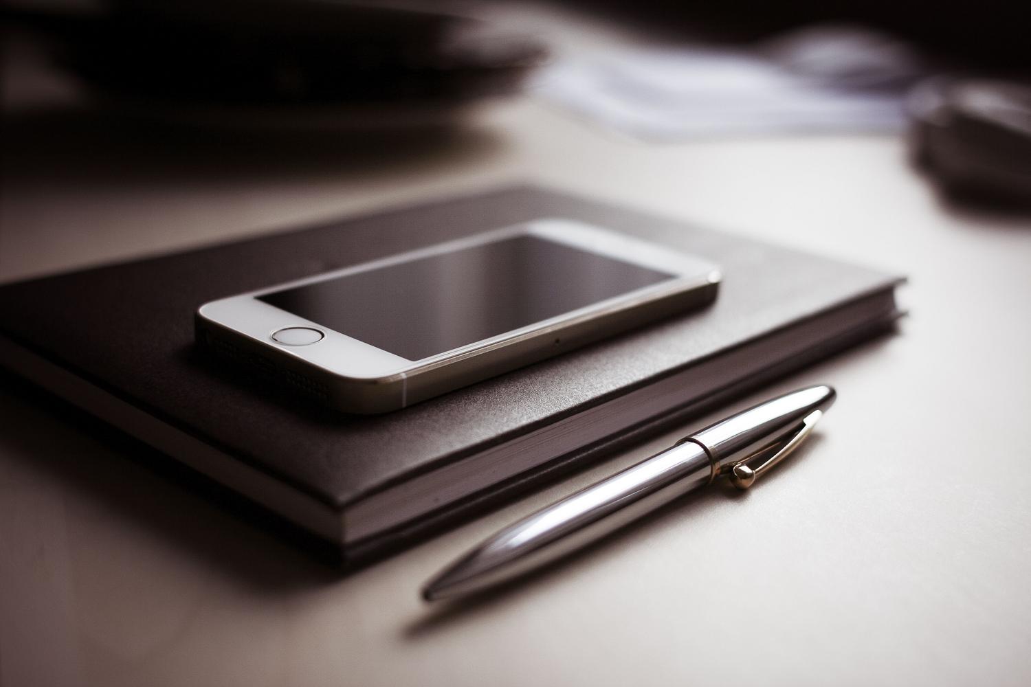 Elegants iPhone uz slēgta piezīmjdatora uz rakstāmgalda, kas simbolizē organizāciju un vadītāja profilu Elegants iPhone uz slēgta piezīmjdatora uz rakstāmgalda, kas simbolizē organizāciju un vadītāja profilu